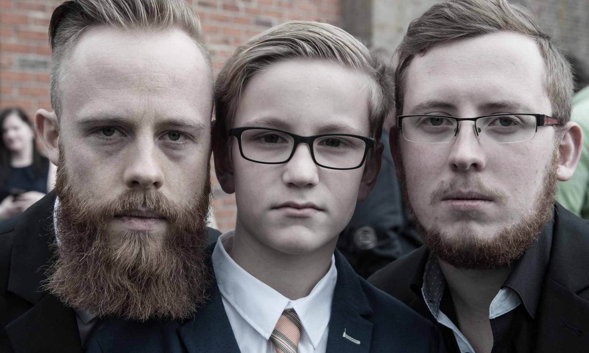 Drei männliche Personen unterschiedlichen Alters blicken ernst in die Kamera – Generationenporträt mit urbanem Hintergrund.